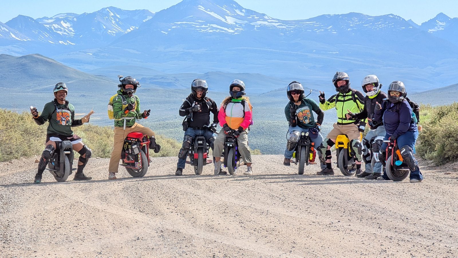 Eight Westwheel riders in helmets sit on their electric unicycles in a line across a gravel road, snow-capped mountains behind them.