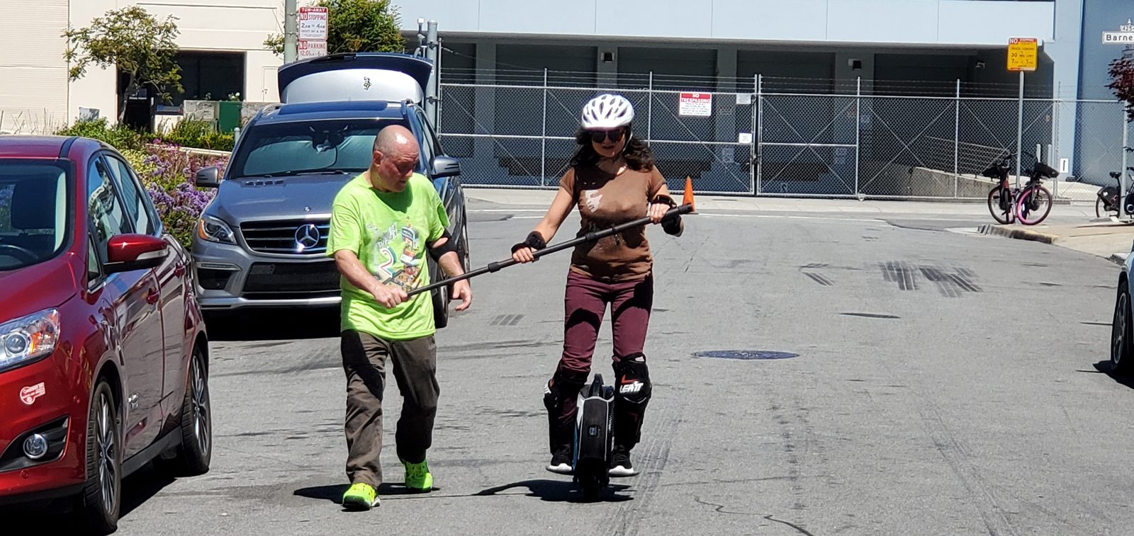 A learner in a white helmet and knee pads rides a single-wheel electric unicycle down a quiet street, both hands on a training bar held by a PEVRA trainer walking beside her.