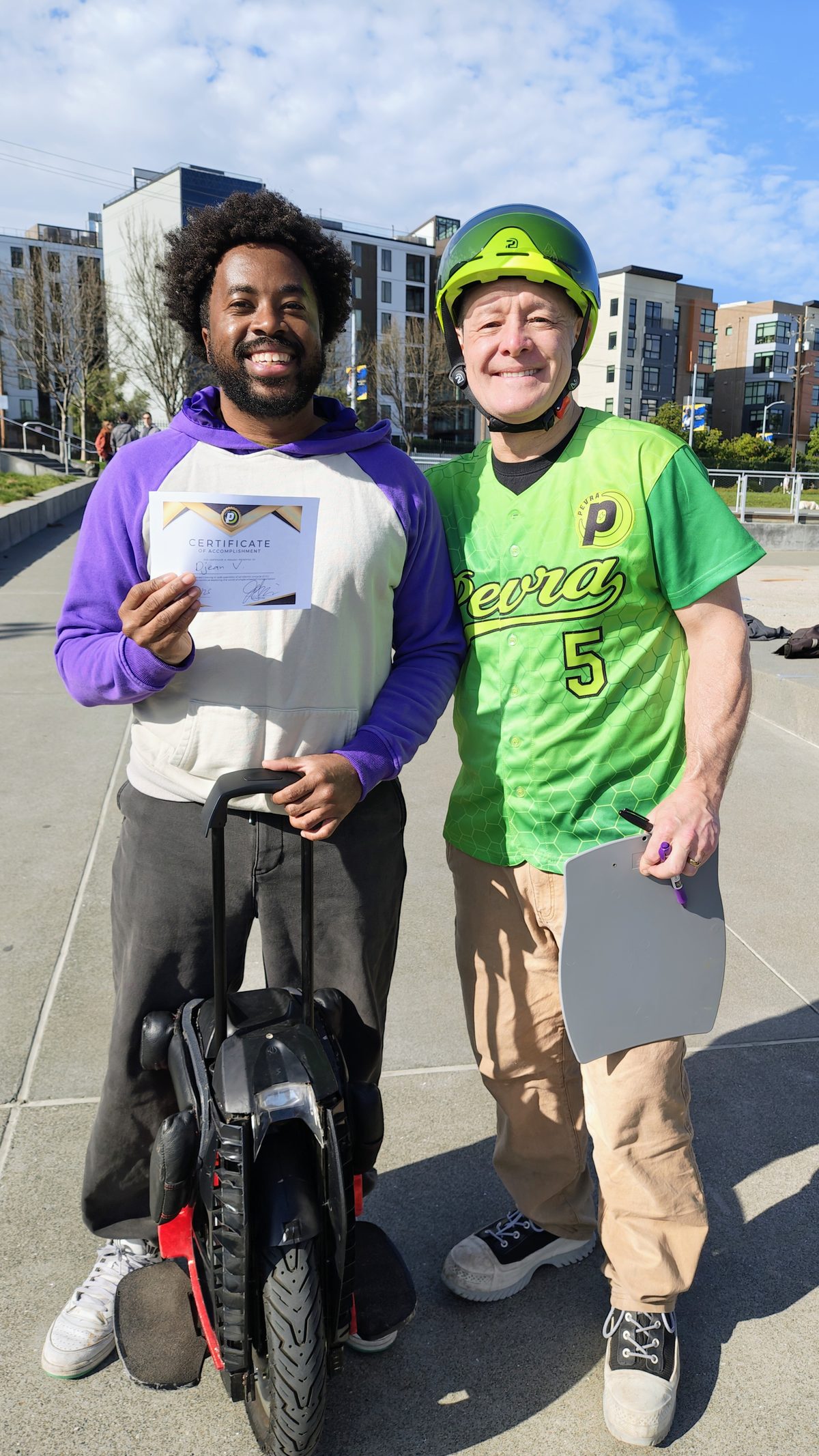 A new rider grins, holding his Certificate of Accomplishment in one hand and his electric unicycle by the trolley handle in the other, beside a PEVRA trainer in a green jersey and helmet.