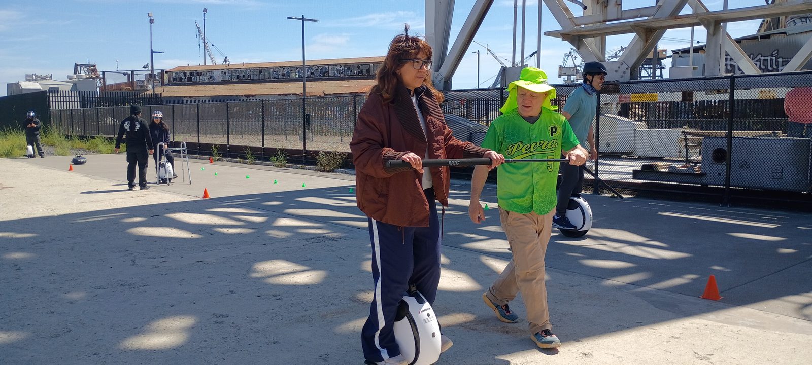 On the Crane Cove waterfront, a learner on a two-wheeled E20 holds a training bar with a PEVRA trainer in a green jersey and sun hat; more learners and cones behind them.