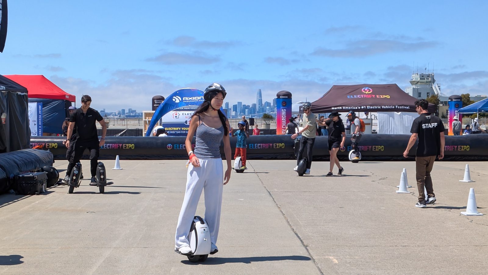 A first-time rider in a helmet glides across a sunny plaza on a two-wheeled Inmotion E20, cones and other riders behind her.