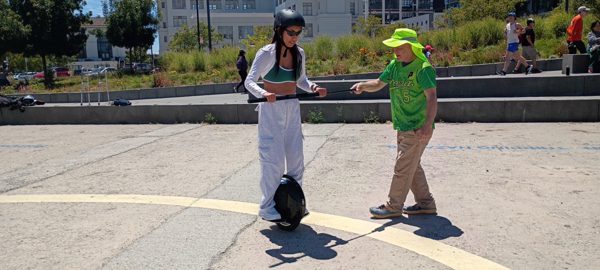 A first-time rider in a helmet balances on an electric unicycle, holding a training bar steadied by a PEVRA instructor in a green jersey, on a sunny San Francisco plaza.