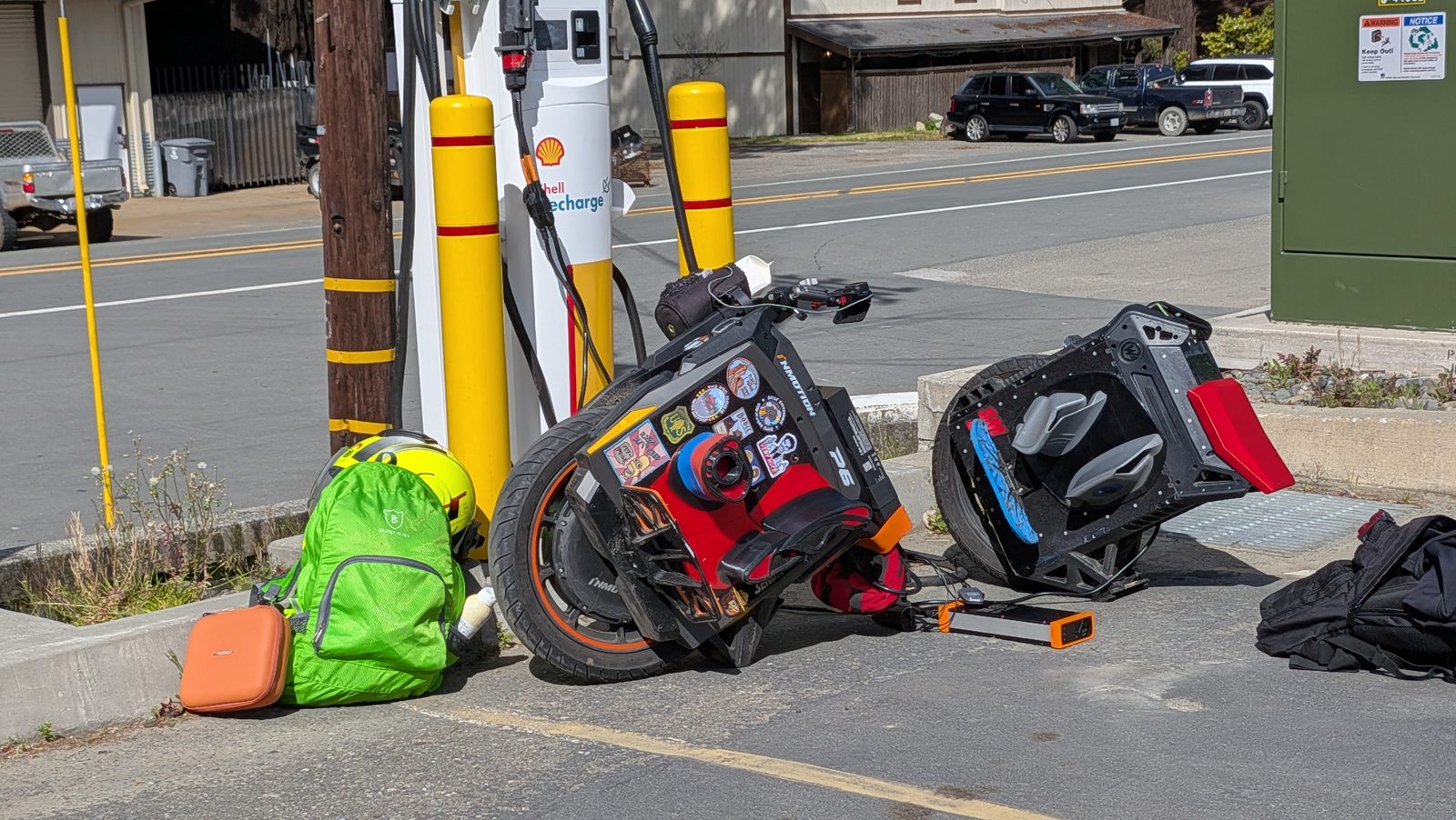 Two electric unicycles lean against the curb at a roadside EV charging post, cables running through an adapter box, with a helmet and packs beside them.