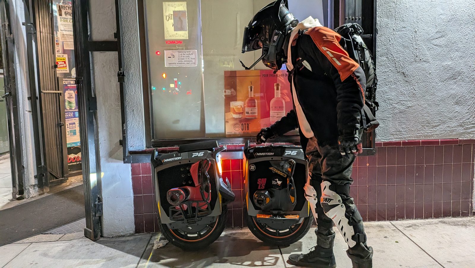 A rider in a full-face helmet, armoured jacket, gloves and knee braces checks two electric unicycles parked against a wall at night.