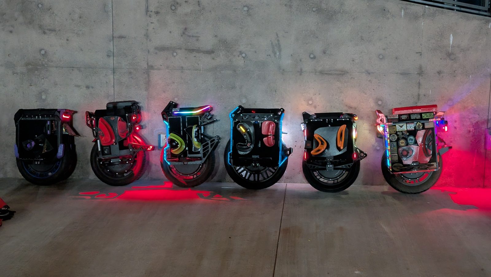 Six electric unicycles of different sizes lined against a concrete wall at night, their trim lights glowing.