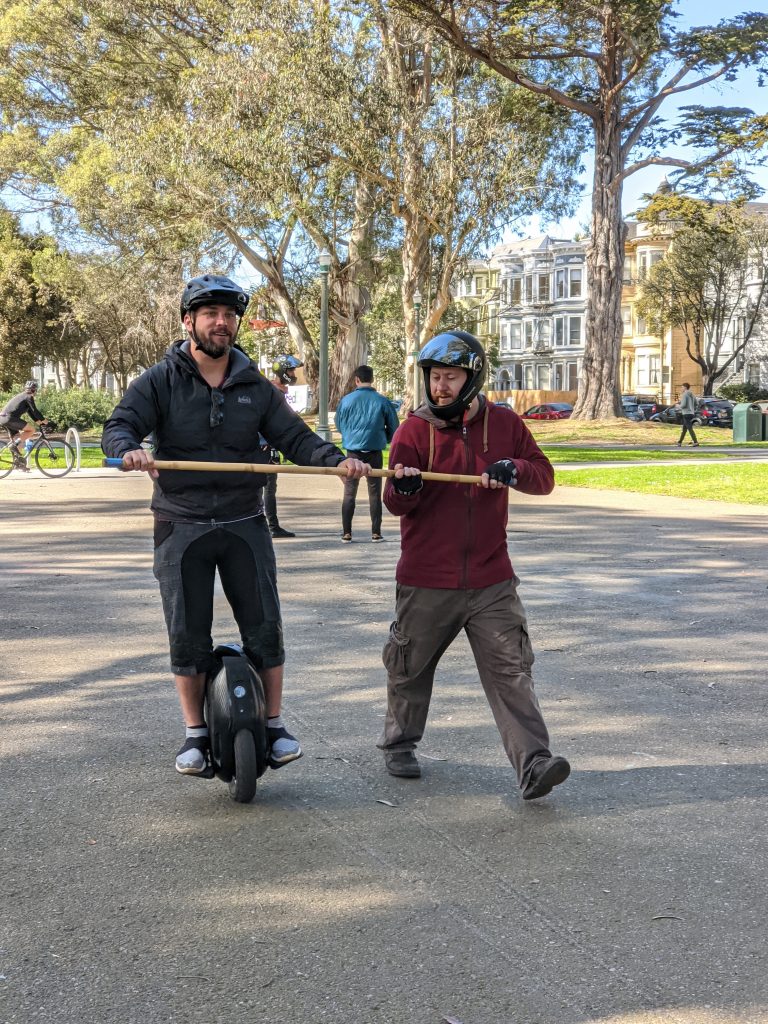 Two people in helmets on a park path: a learner balancing on an electric unicycle with both hands on a long wooden training bar, and a trainer crouched at the far end of the same bar, holding it steady.