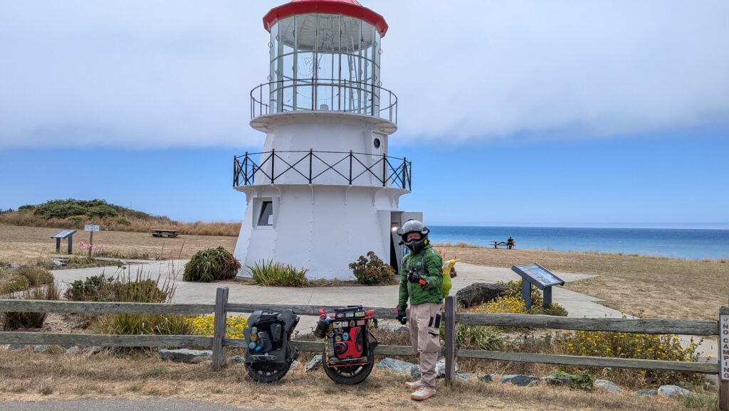 A rider in a full-face helmet standing with two loaded wheels at the Cape Mendocino lighthouse.